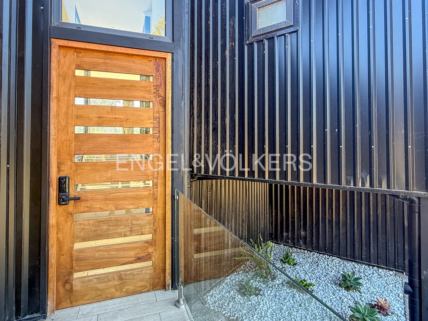 Exterior shot of a modern home with a wooden door with horizontal glass panels, black corrugated metal siding, and a small rock garden.