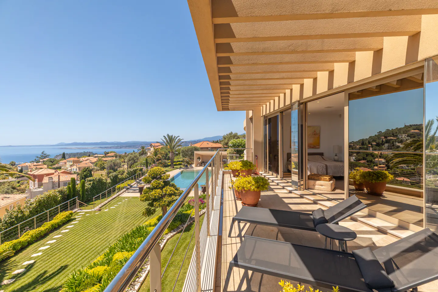 View from a tan balcony with lounge chairs, potted plants, and a stainless steel railing overlooking a pool, lawn, and ocean view.