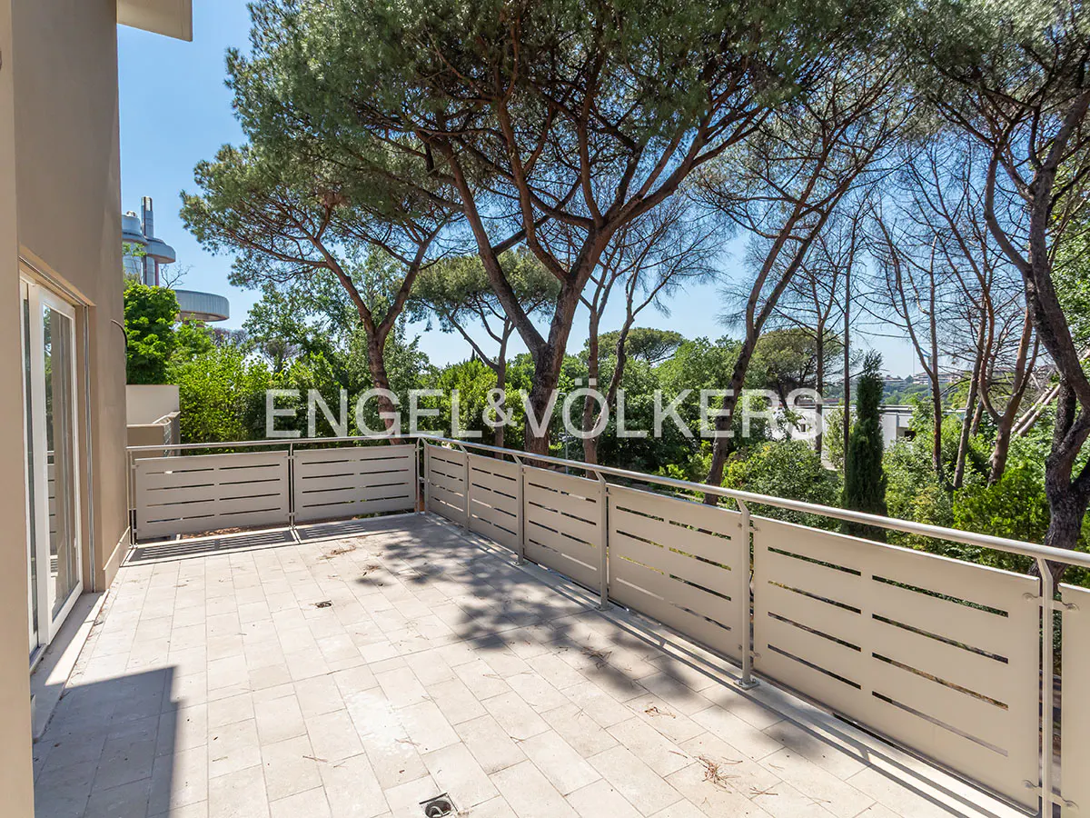 Spacious tiled balcony with modern beige railings overlooking a lush green garden with tall pine trees under a blue sky.