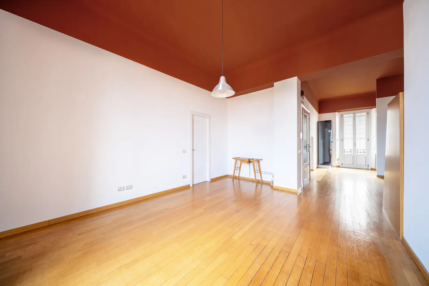 Bright, empty room with hardwood floors, white walls, and a brown ceiling. A pendant light hangs above a small wooden table.