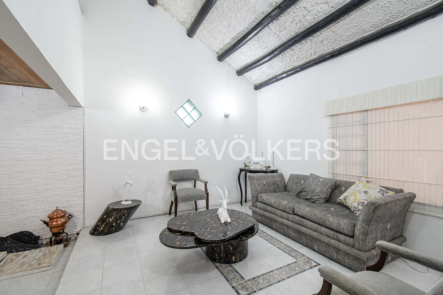 Living room with gray sofa, chairs, and unique tables. White walls, textured ceiling with dark beams, and tiled floor. Natural light from window.