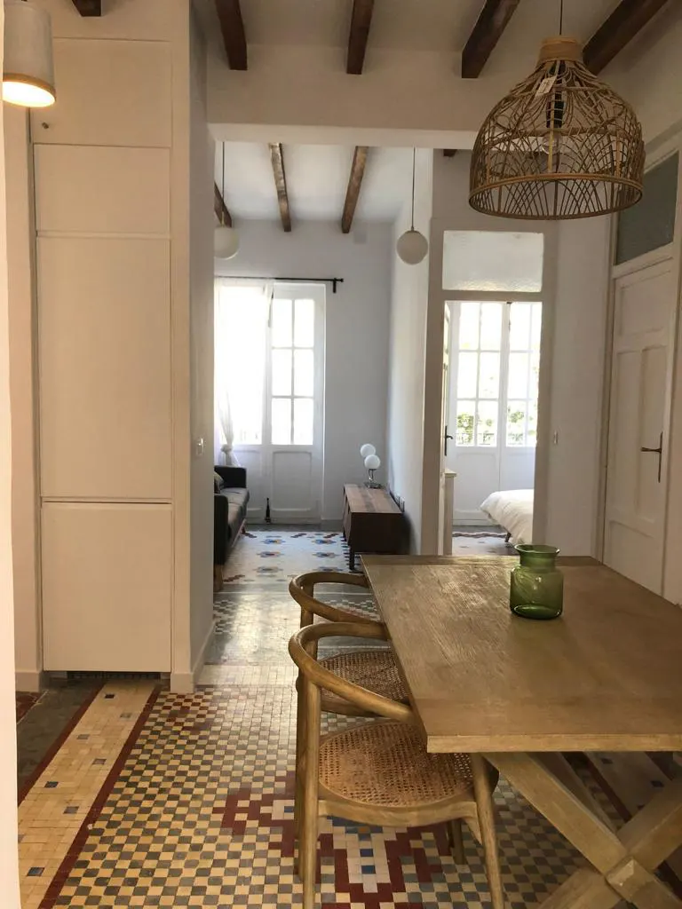 Interior view of a bright, open-concept home with a wooden dining table, rattan chairs, and colorful tiled floors. Living and bedroom areas visible.