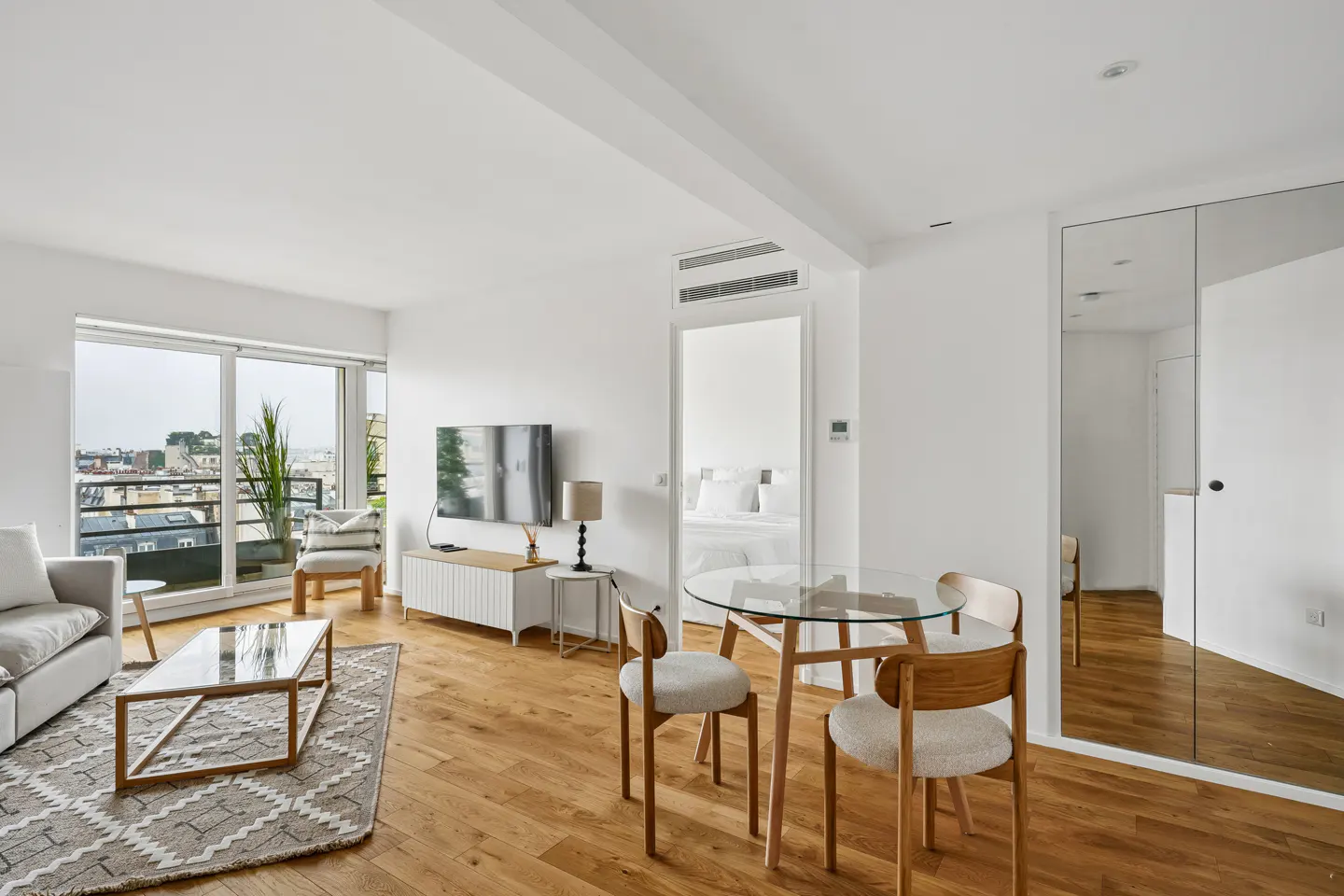 Bright, modern apartment interior with wood floors, a glass dining table, and a view of the city skyline.