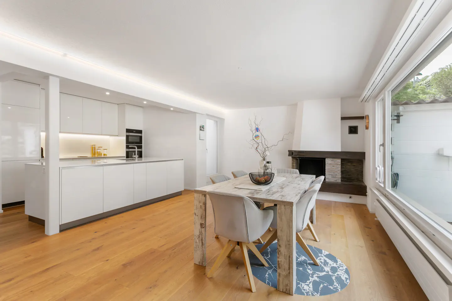 Bright, modern kitchen and dining area with white cabinets, wood floors, a stone fireplace, and a large window.