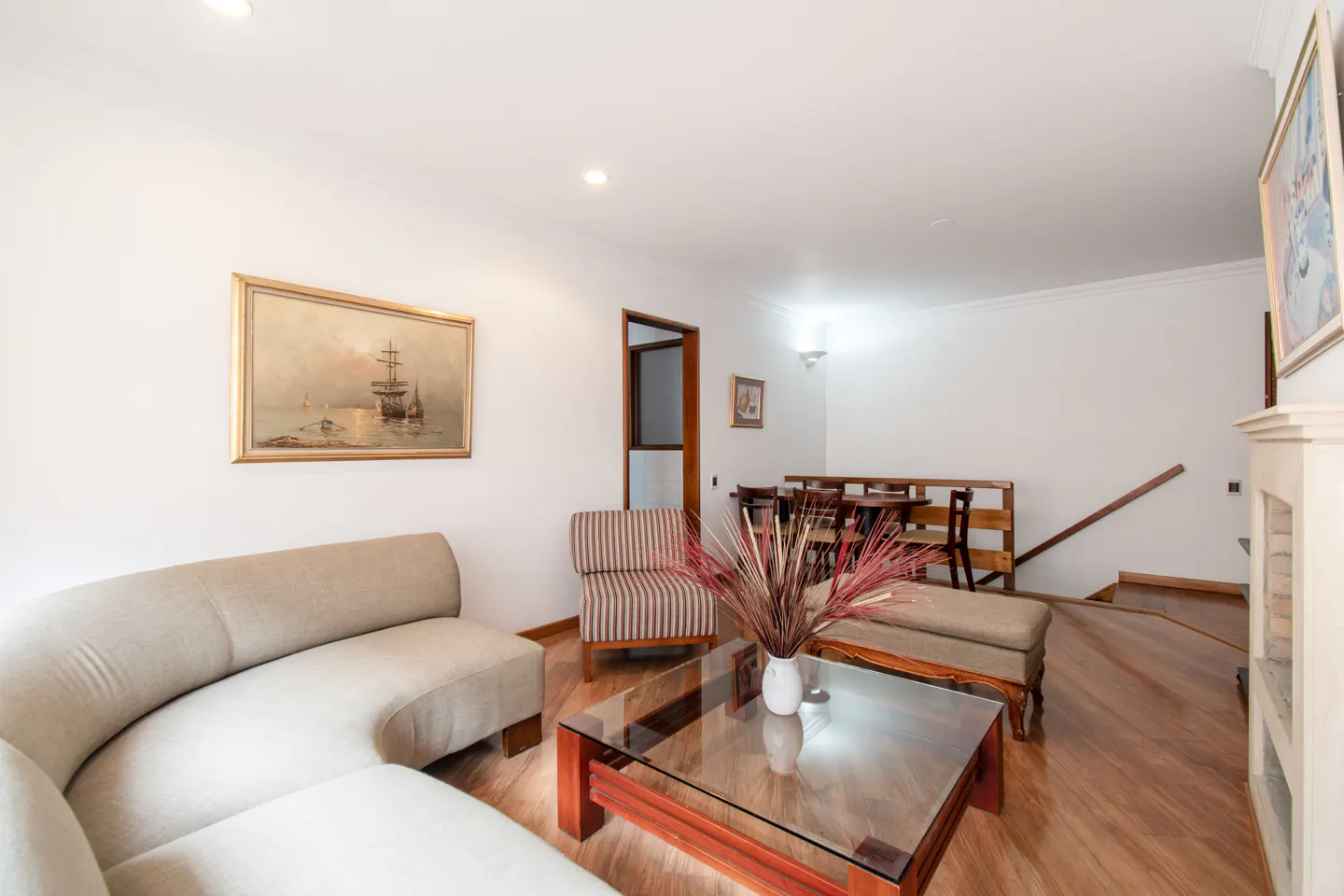 Living room with beige sofa, striped chair, glass coffee table, and wood floors. A painting of a ship hangs on the white wall.