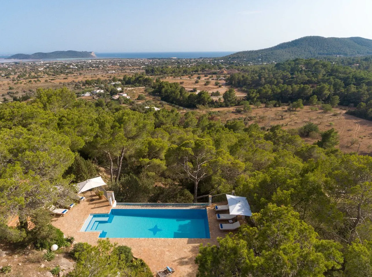 Aerial view of a blue swimming pool surrounded by trees, lounge chairs, and umbrellas, with a view of the ocean in the background.