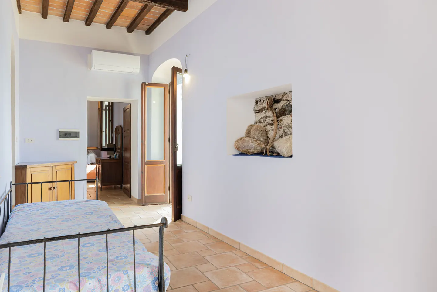 Bedroom with twin beds, terracotta tile floor, and white walls. A niche displays rocks and a branch. Open door leads to another room.