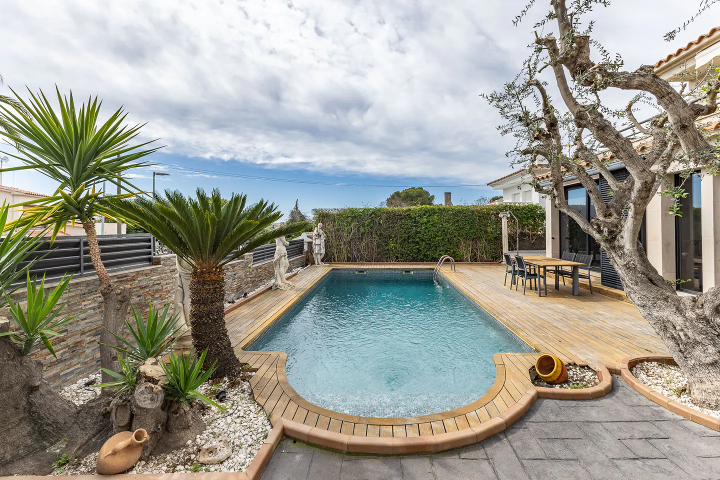 Backyard view of a rectangular pool with a wooden deck, statues, and lush greenery. A table and chairs sit near the house.