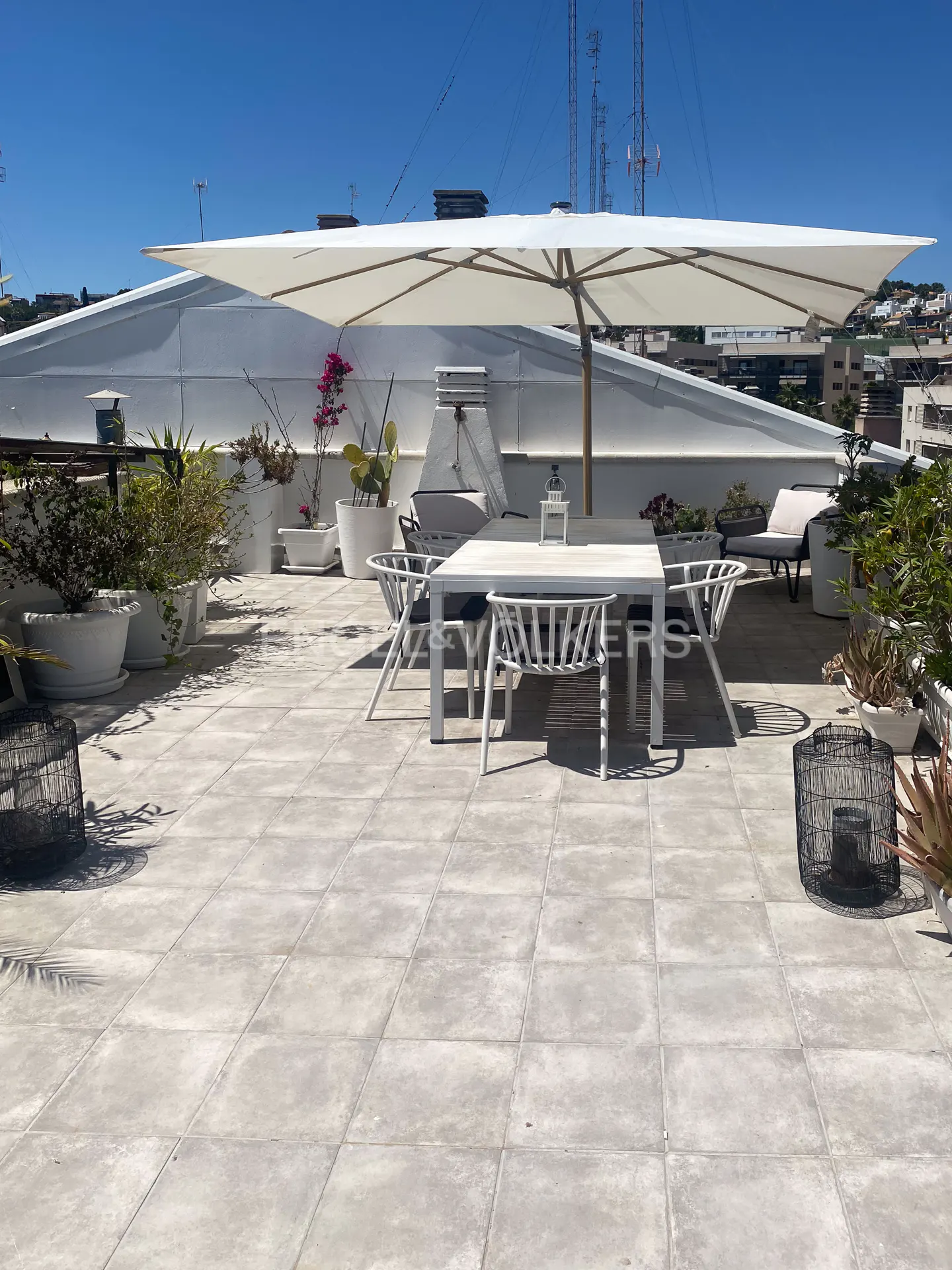 Rooftop patio with white table, chairs, and large umbrella. Potted plants line the perimeter, with city buildings in the background.