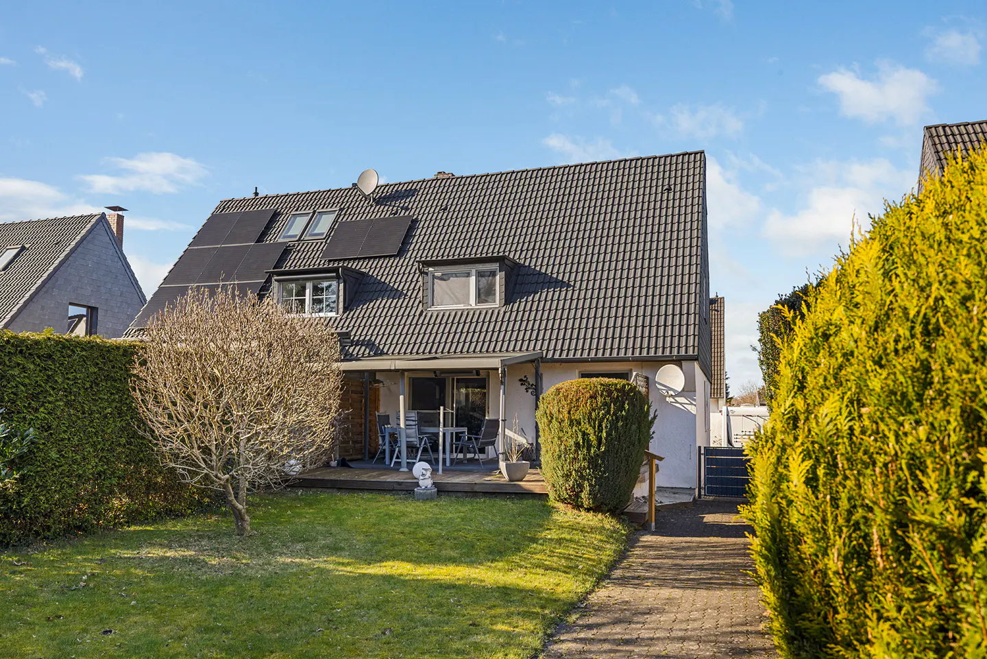 A two-story house with a gray tiled roof and solar panels, a patio with furniture, and a green lawn.