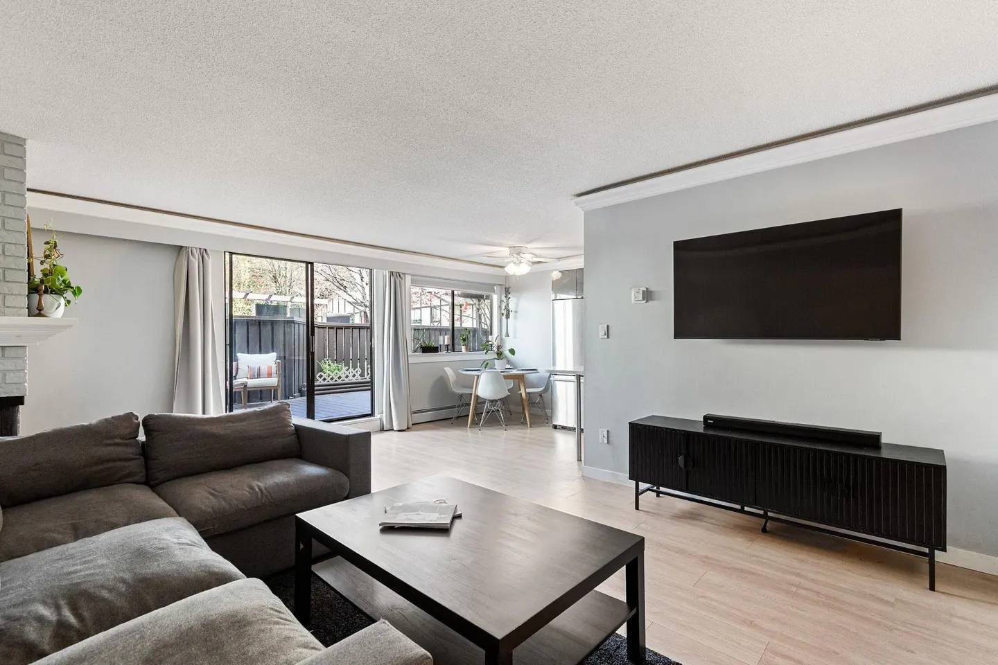 Modern living room with a grey sectional, dark coffee table, wall-mounted TV, and a dining area with sliding glass doors.