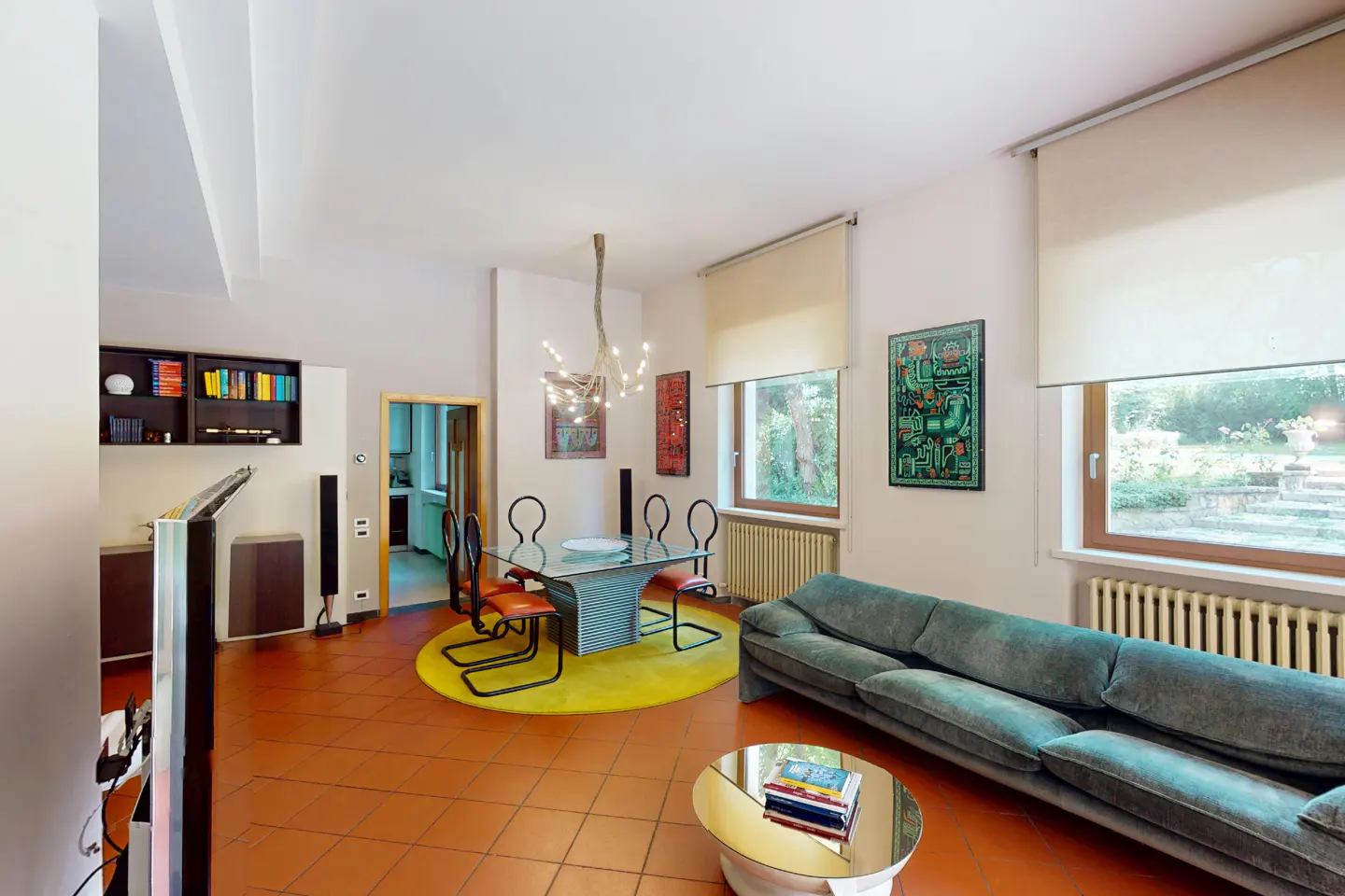 Bright living room with terracotta tile floor, blue sofa, glass table, and yellow rug. Windows show a garden view.
