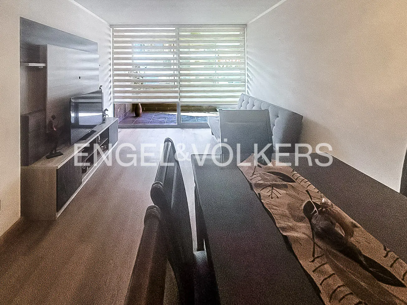Living room with a gray sofa, TV, and a dark wood table with a brown runner. Blinds cover the sliding glass doors.