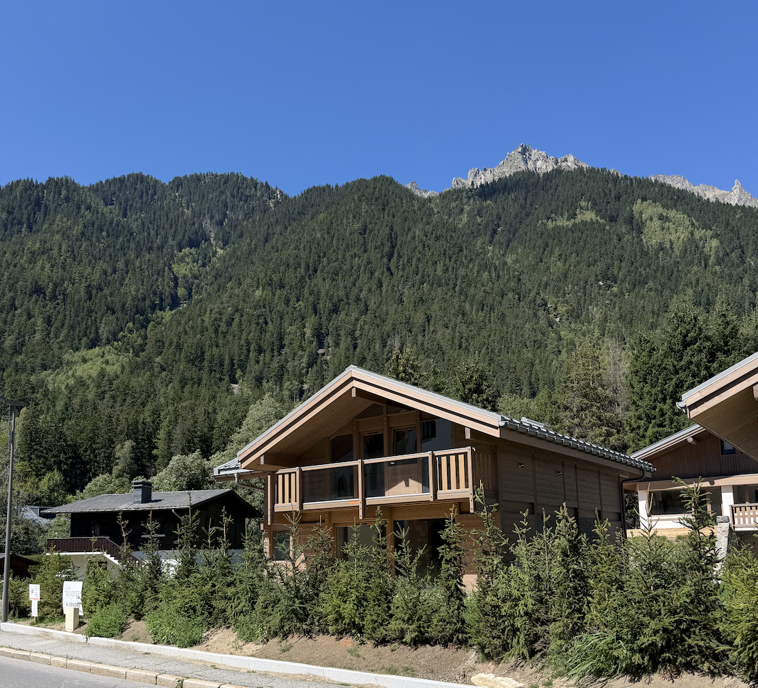 Modern wooden chalet with a balcony, nestled at the base of a lush, pine-covered mountain under a clear blue sky. [AI-generated text]