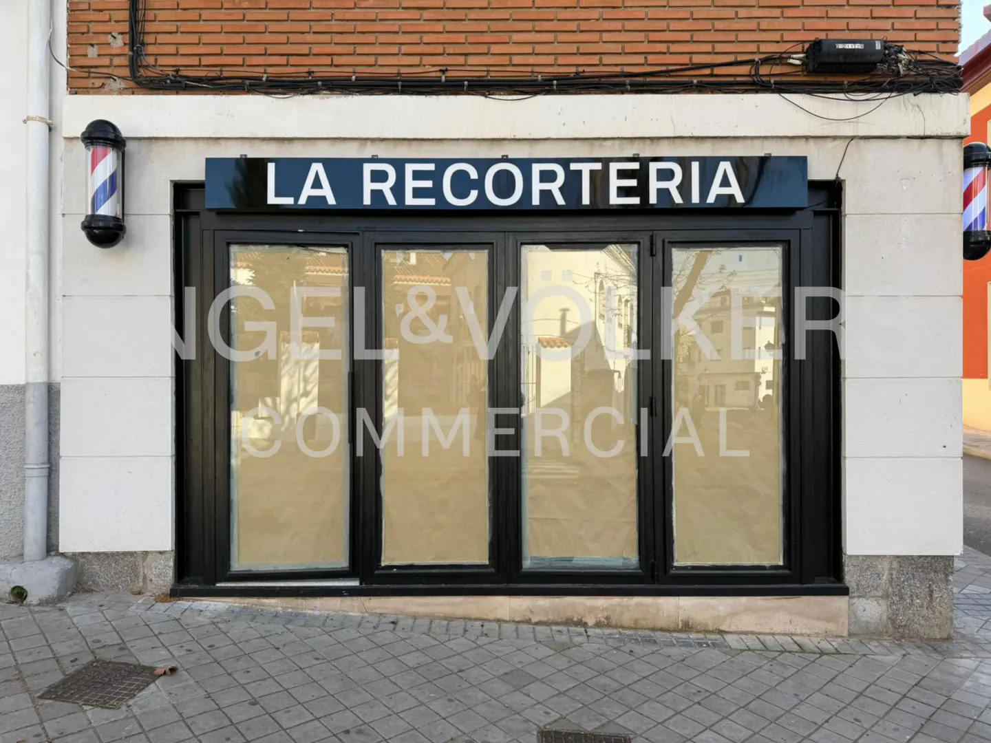 Exterior of "La Recorteria" barber shop with Engel & Volkers Commercial sign on the windows. Barber poles flank the black-framed glass doors.