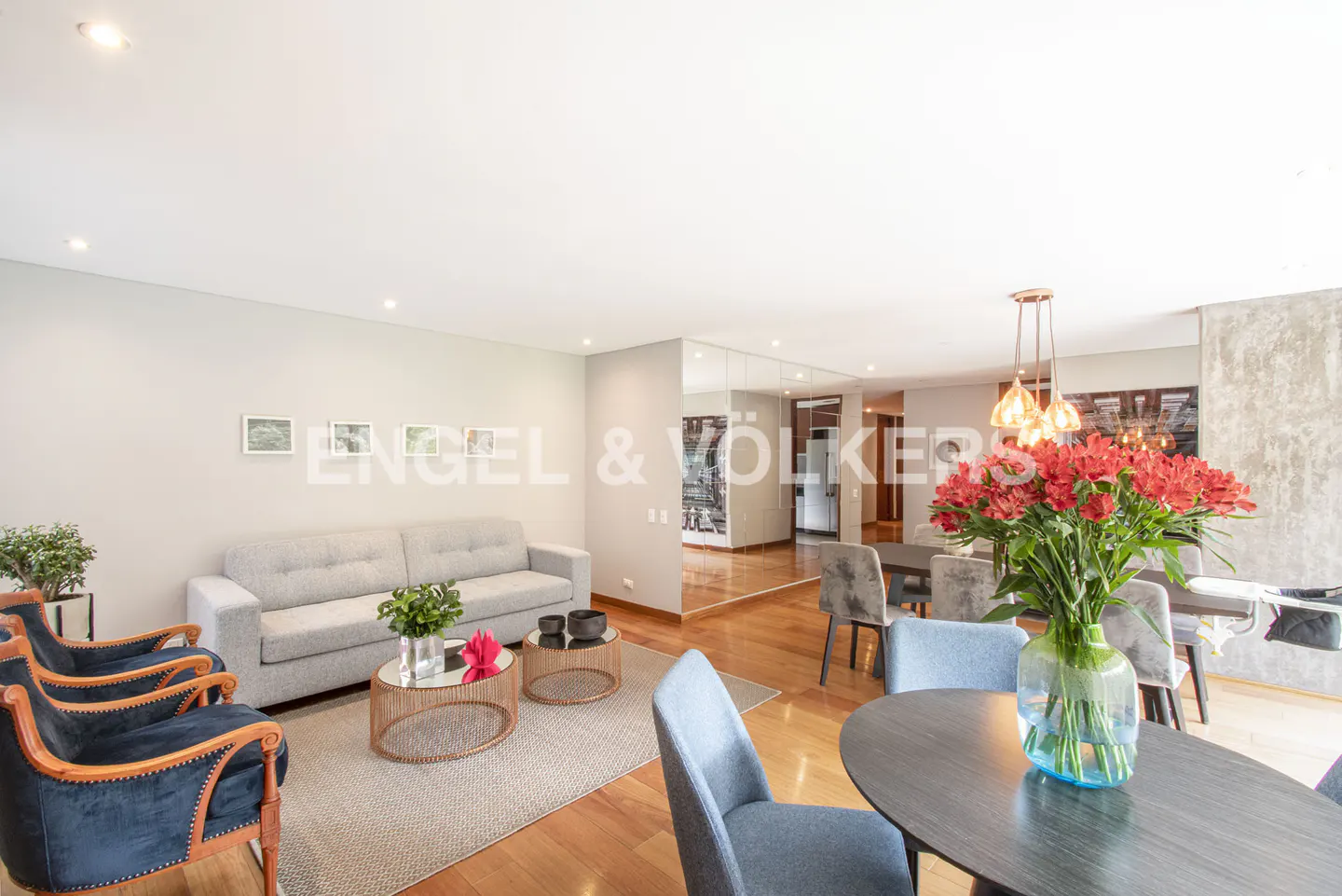 Open-concept living and dining area with a gray sofa, blue armchairs, and a round dining table with red flowers.