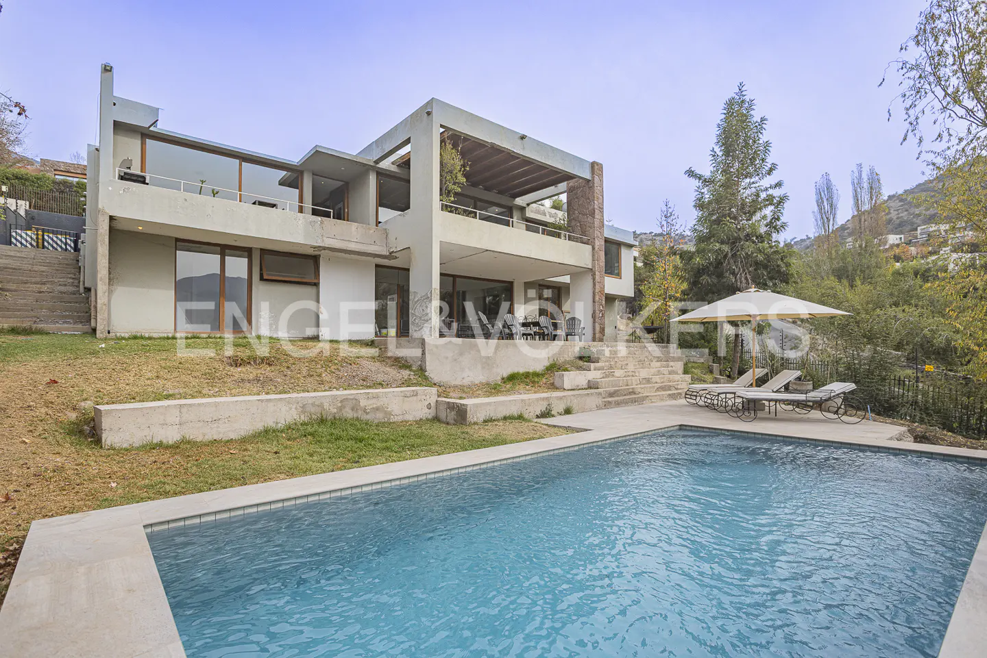 Modern two-story home with a swimming pool, lounge chairs, umbrella, and terraced backyard on a sunny day.