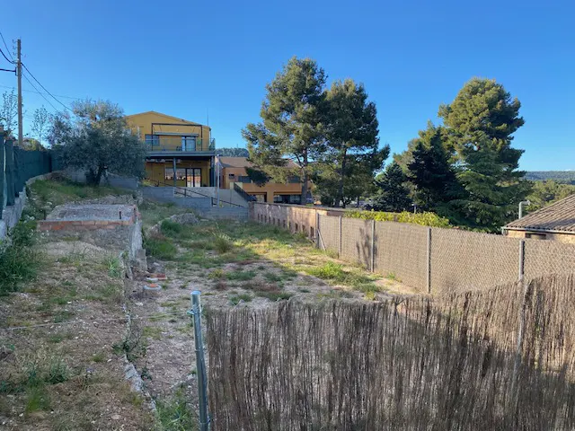 Vacant lot with a straw fence, green grass, and a yellow house in the background under a clear blue sky.