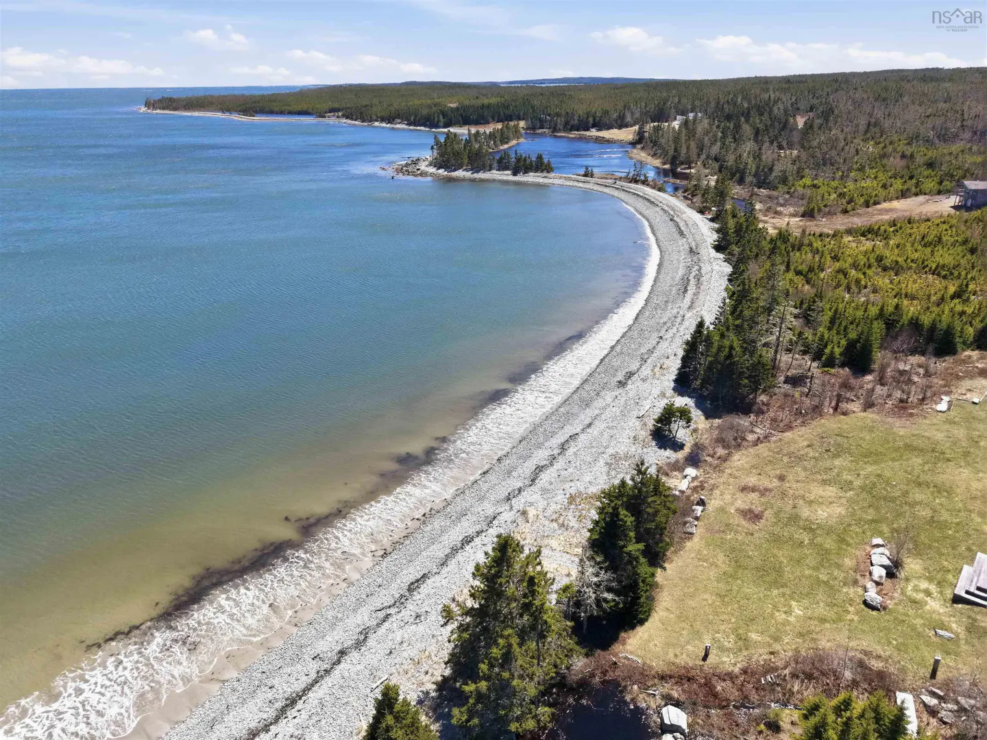 Aerial view of a curved rocky beach meeting blue ocean water, bordered by a dense evergreen forest and grassy field.