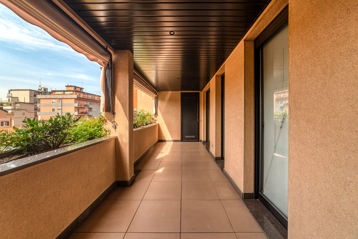 A long, covered balcony with beige walls, a dark ceiling, and tiled floor. Plants line the outer edge, with a city view in the background.