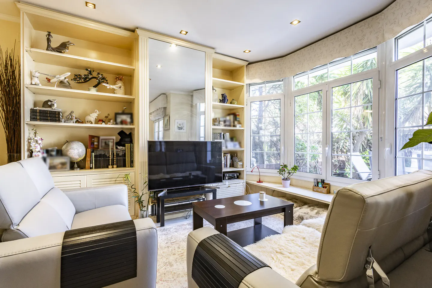 Living room with cream-colored built-in shelves, a large mirror, and a flat-screen TV. Two light gray sofas face each other, with a dark wood coffee table in between. Large windows let in natural light.
