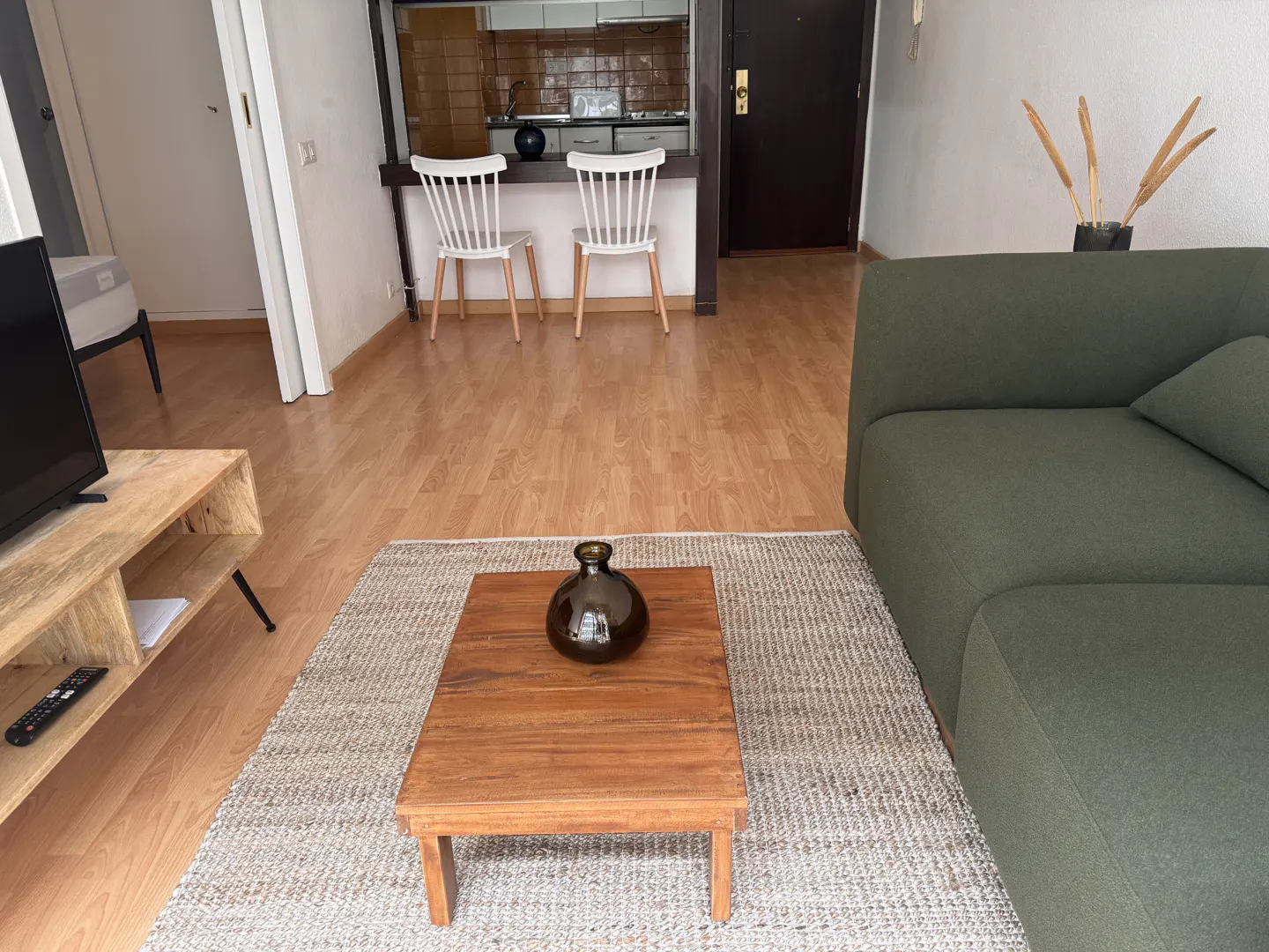Living room with a green sofa, wood coffee table with a vase, and a view of the kitchen with white chairs.