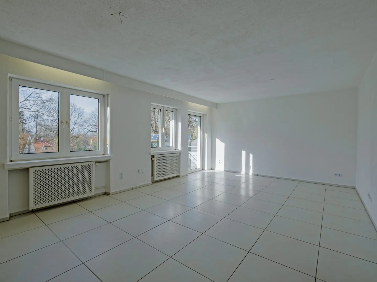 Bright, empty room with white walls and tiled floor. Windows show trees outside. Radiators under windows. Sunlight streams in.