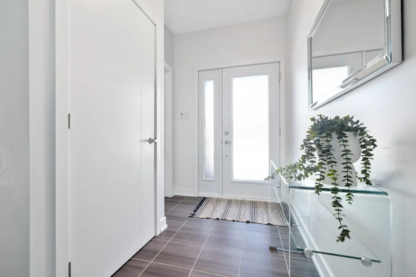 Bright entryway with white door, glass console table with plant, and a mirror. Brown tile floor and a striped rug.