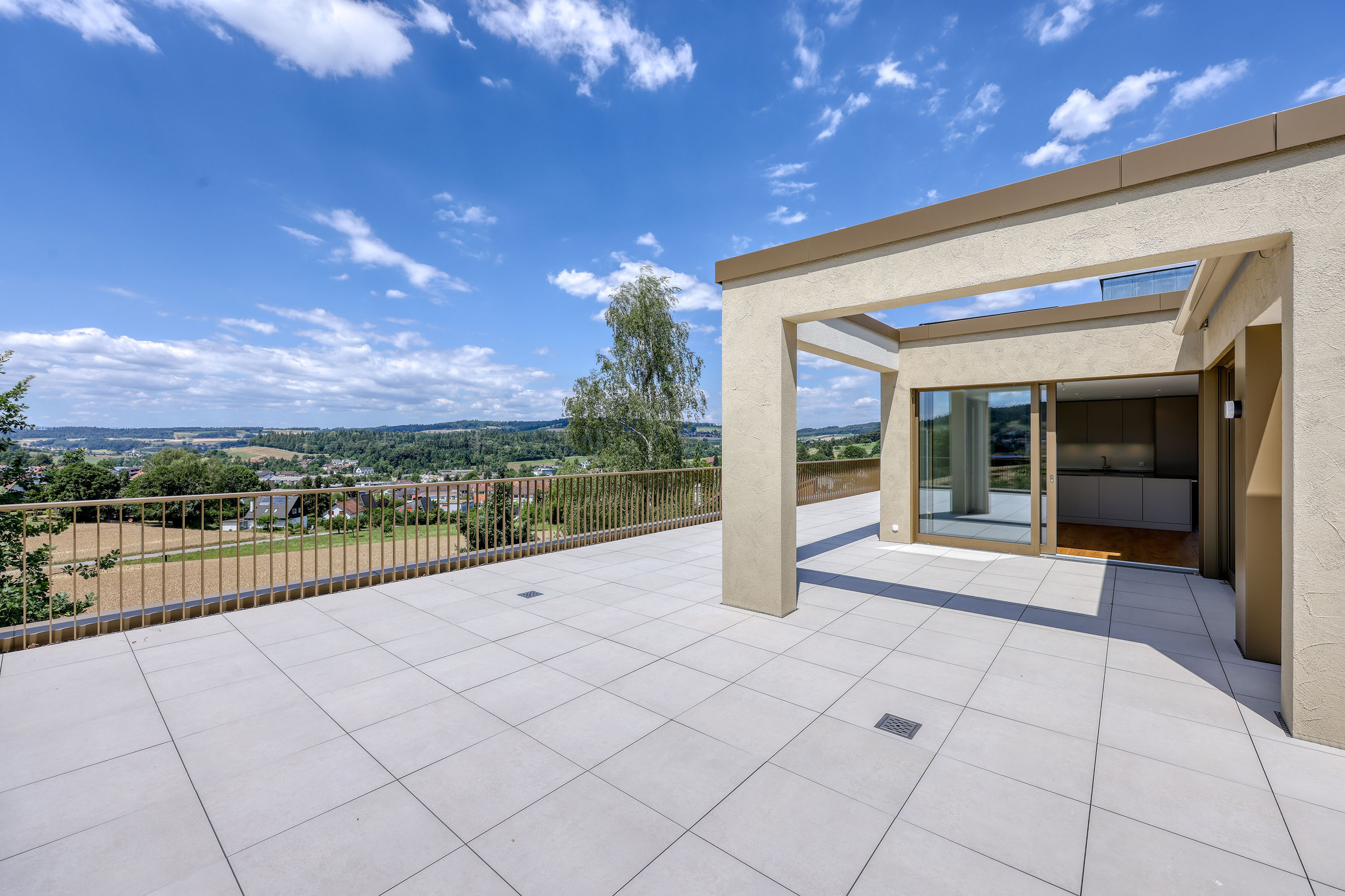 Spacious modern rooftop terrace with light-colored tiles, metal railing, and a scenic view of a town under a blue sky. [AI-generated text]