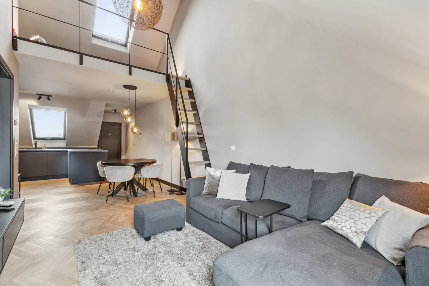 Living room with gray sectional sofa, ottoman, and rug. A black staircase leads to a loft with skylights. Kitchen and dining area in the background.