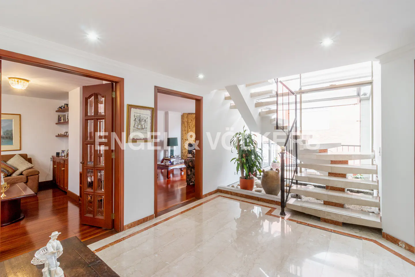 Bright foyer with marble floors, wood trim, and open staircase. Living room visible through doorway. Plant near stairs.