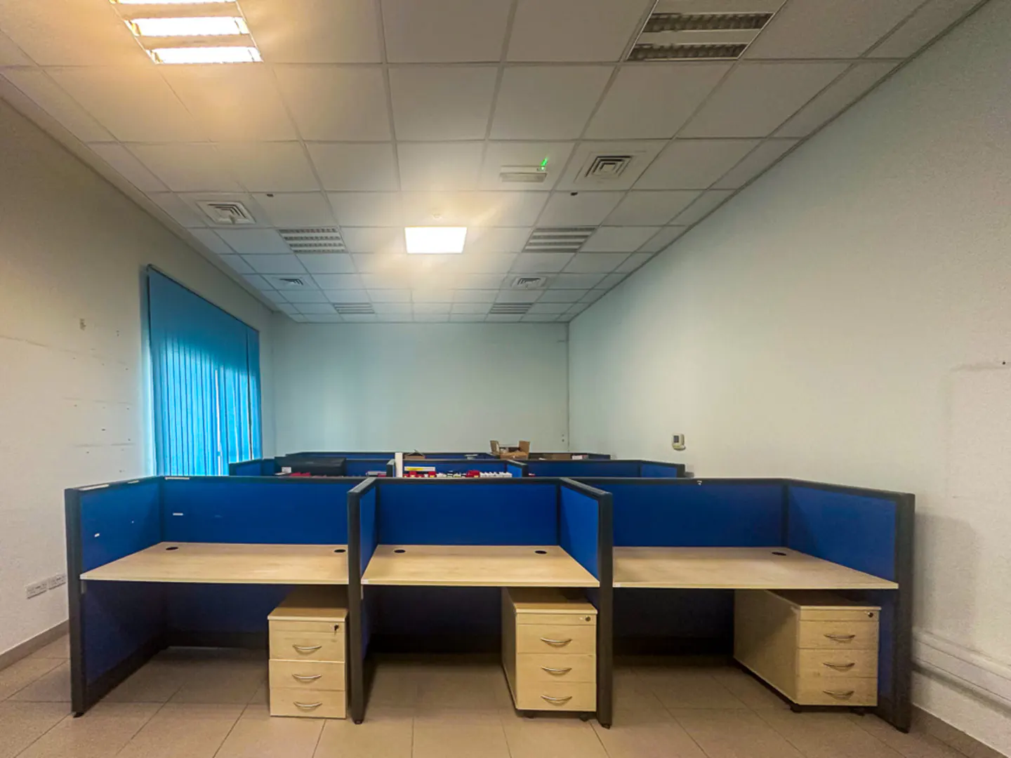 Empty office space with blue cubicles and beige desks. White walls and ceiling with fluorescent lighting. Blue curtains on the left.