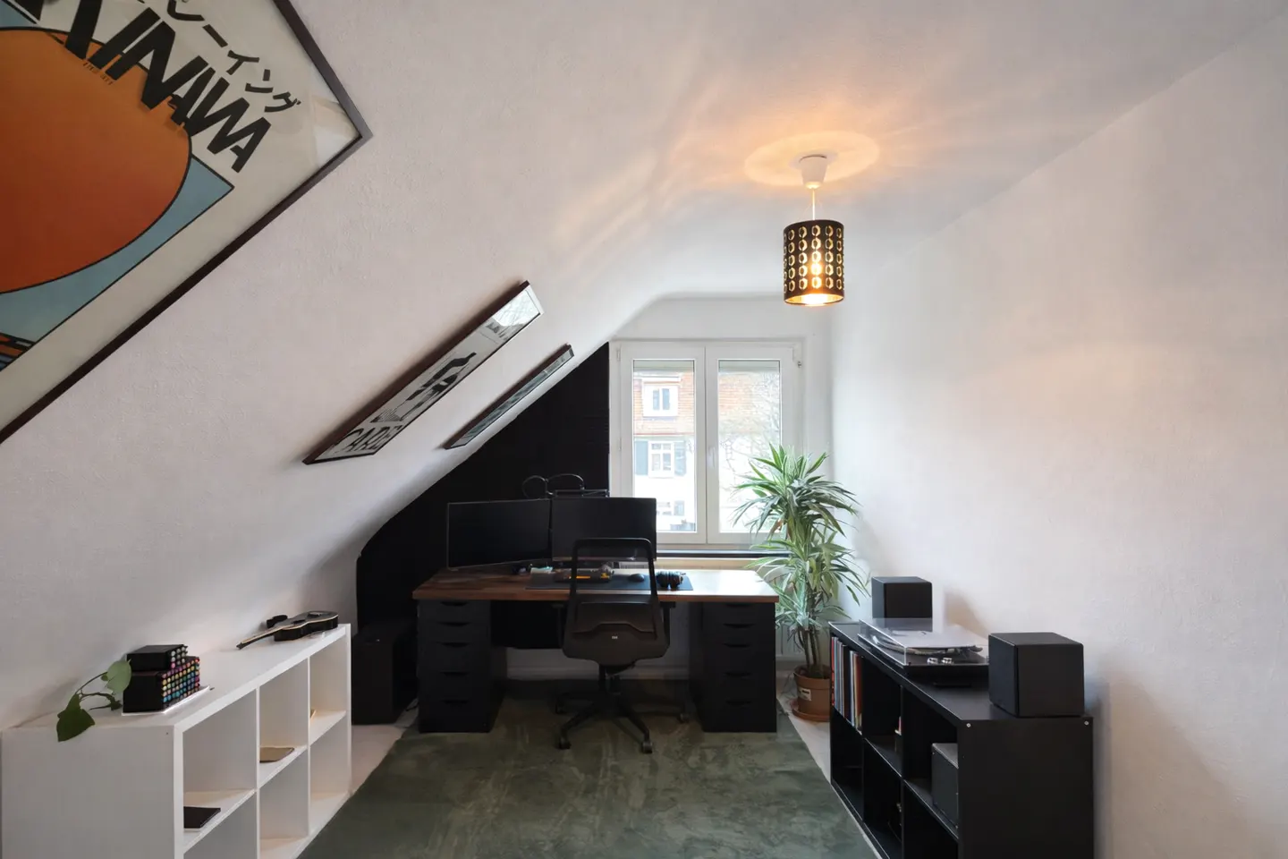 Attic office with white walls, a black accent wall, and a window. A desk with two monitors and a chair sit on a green rug. Shelves line the walls.