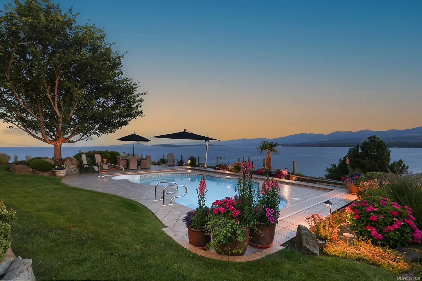 Outdoor pool at dusk with ocean view. Patio furniture, umbrellas, and potted flowers surround the pool.