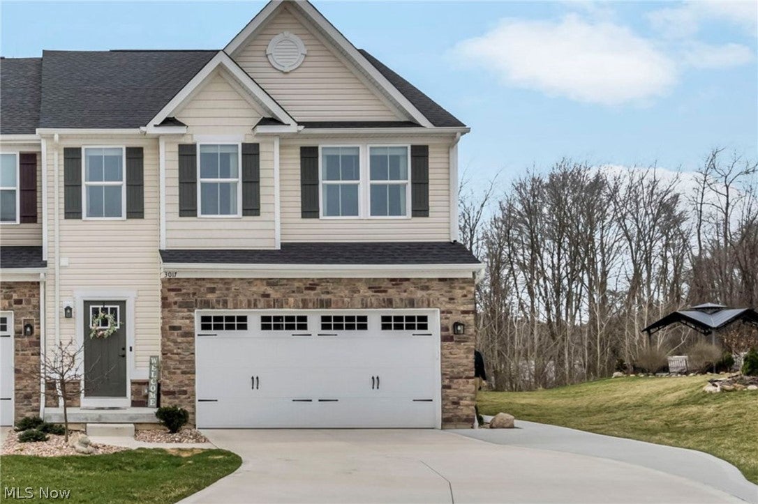 Two-story townhouse with beige siding, stone facade, white garage door, and a dark grey roof under a blue sky. [AI-generated text]