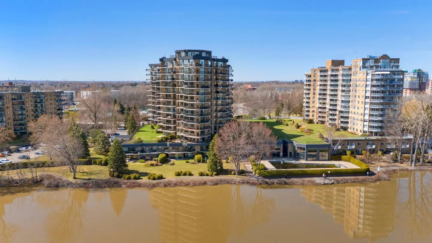 Aerial view of modern condo buildings with balconies, green roofs, and a pool by a brown river under a blue sky.