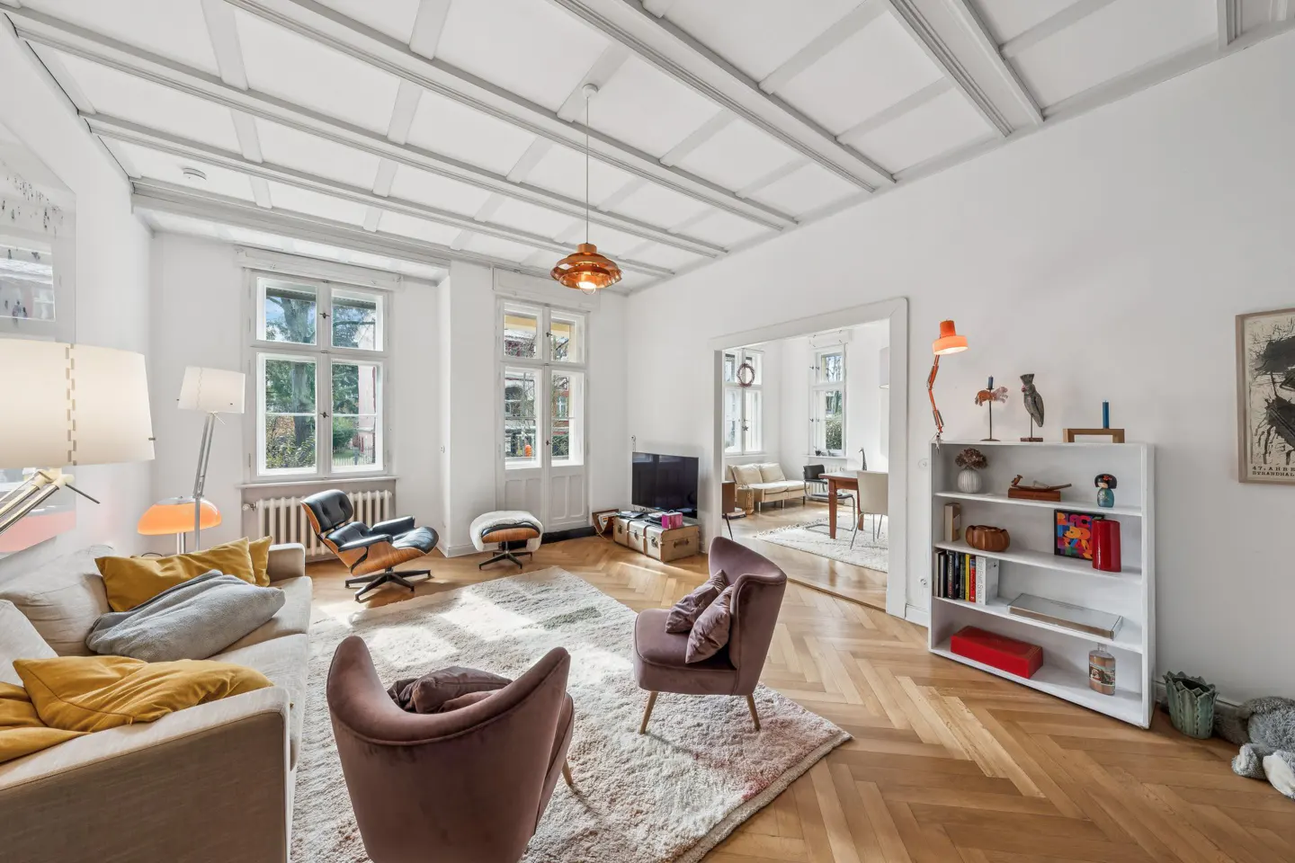 Bright living room with white walls, herringbone wood floors, and a coffered ceiling. Two mauve chairs face a sofa with yellow pillows.