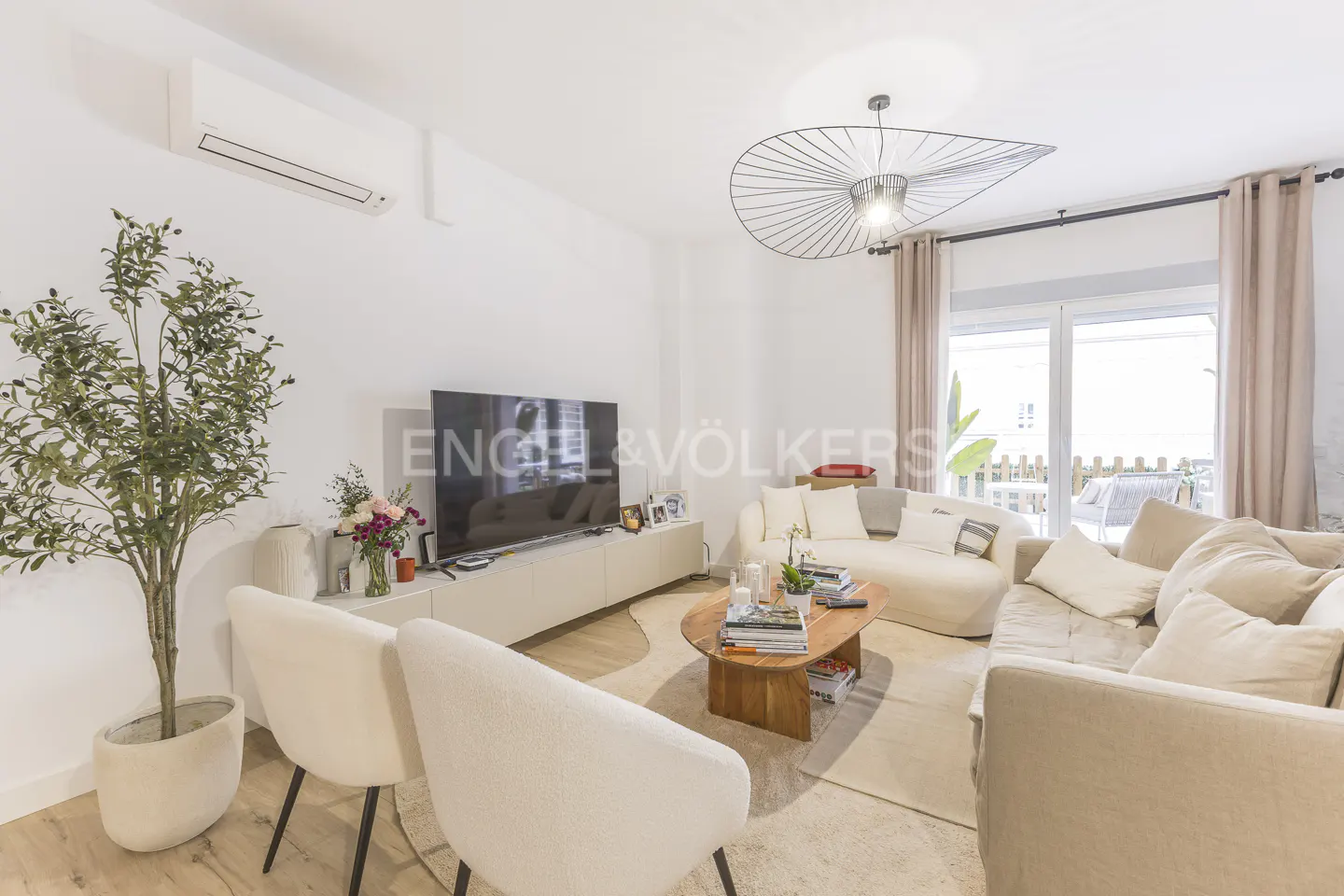 Bright living room with white walls, beige sofas, a wooden coffee table, and a large TV. A potted plant sits near two white chairs.