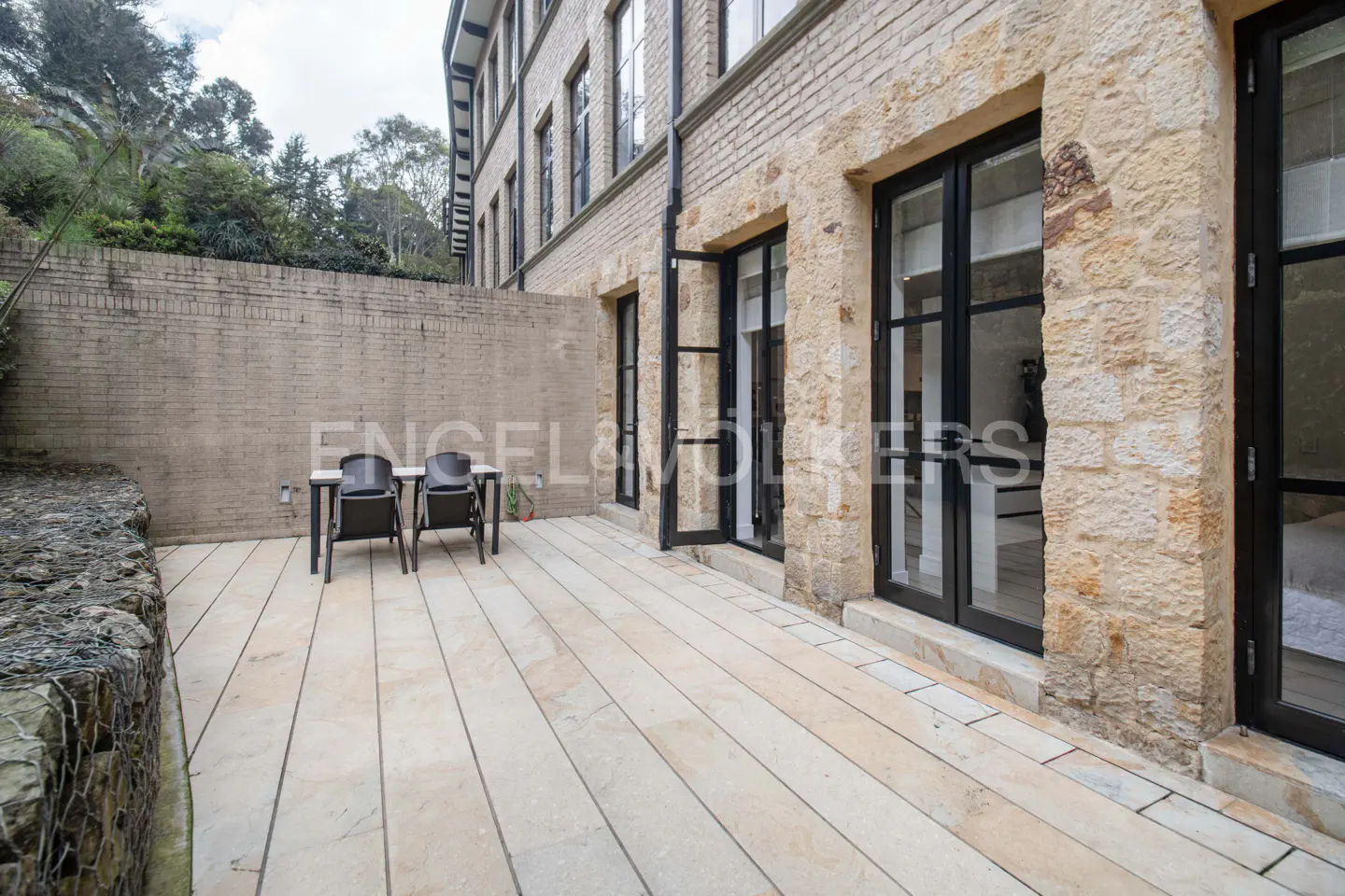 Outdoor patio with stone flooring, a table and two chairs, and a stone building with black framed windows.