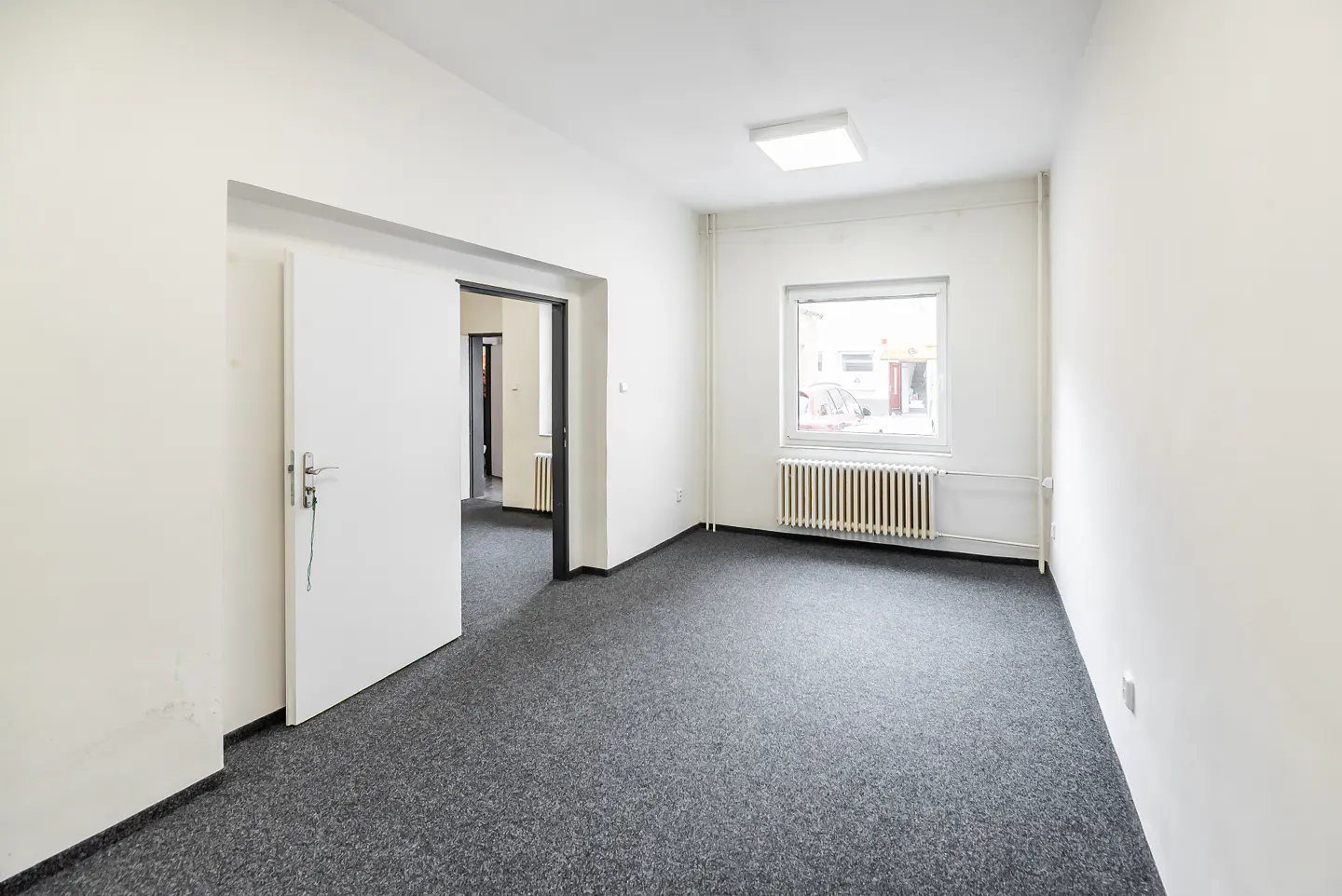 Bright, empty room with white walls, gray carpet, and a window. A door is ajar, revealing another room. Radiator under the window.