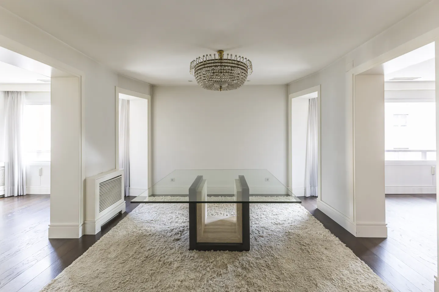 Bright dining room with a glass table on a shag rug under a crystal chandelier. White walls and dark wood floors.
