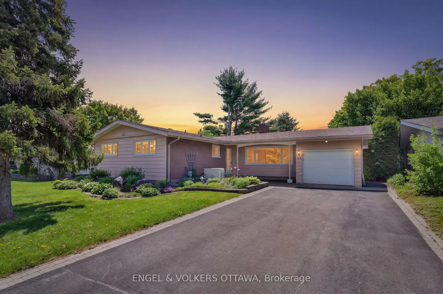A single-story pink house with a garage, green lawn, and trees under a colorful sunset sky.