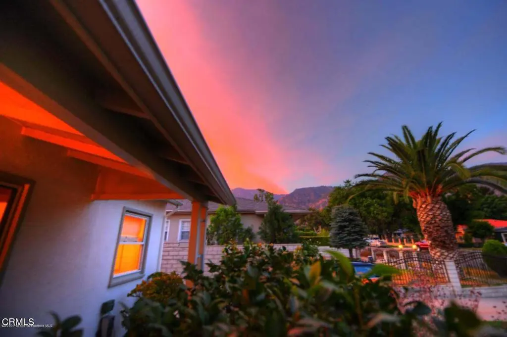A house exterior at dusk with a vibrant pink and blue sunset over distant mountains, a palm tree, and glowing windows.