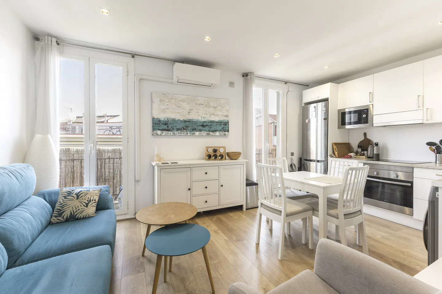 Bright apartment interior with a blue sofa, white kitchen cabinets, and a dining table. Balcony doors open to a cityscape.