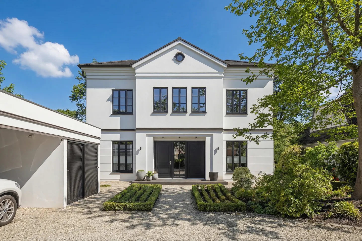 Two-story white house with black window frames and doors, a gravel driveway, and green landscaping on a sunny day.
