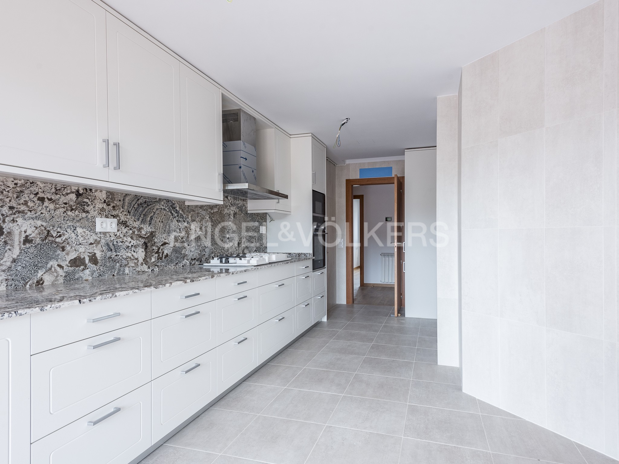 Modern kitchen with white cabinets, granite countertops, and grey tiled floors, leading to an open doorway.