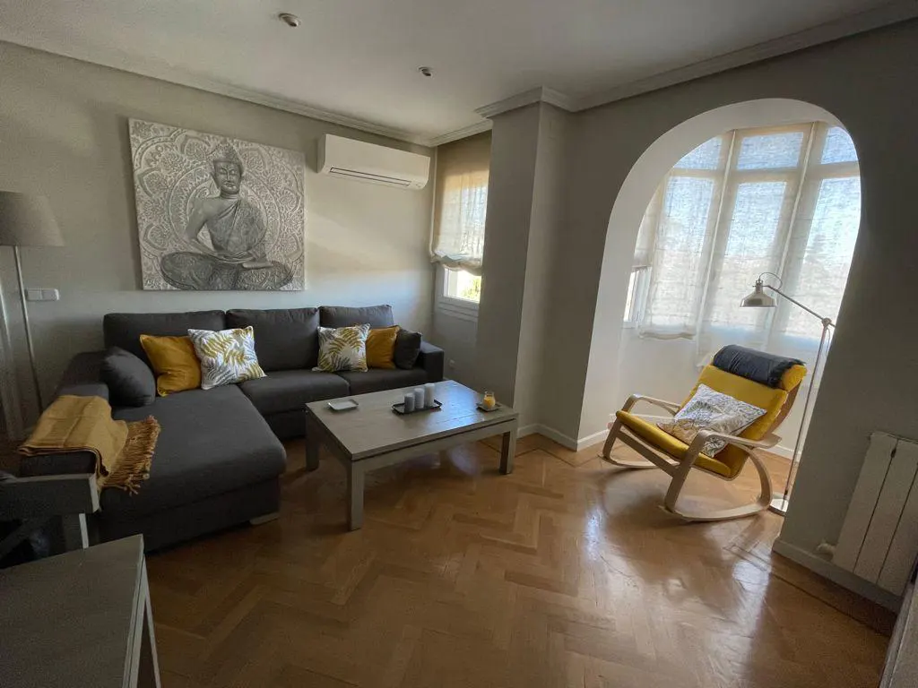 Living room with gray sofa, yellow pillows, and Buddha art. A yellow rocking chair sits in an arched window nook. Hardwood floors.