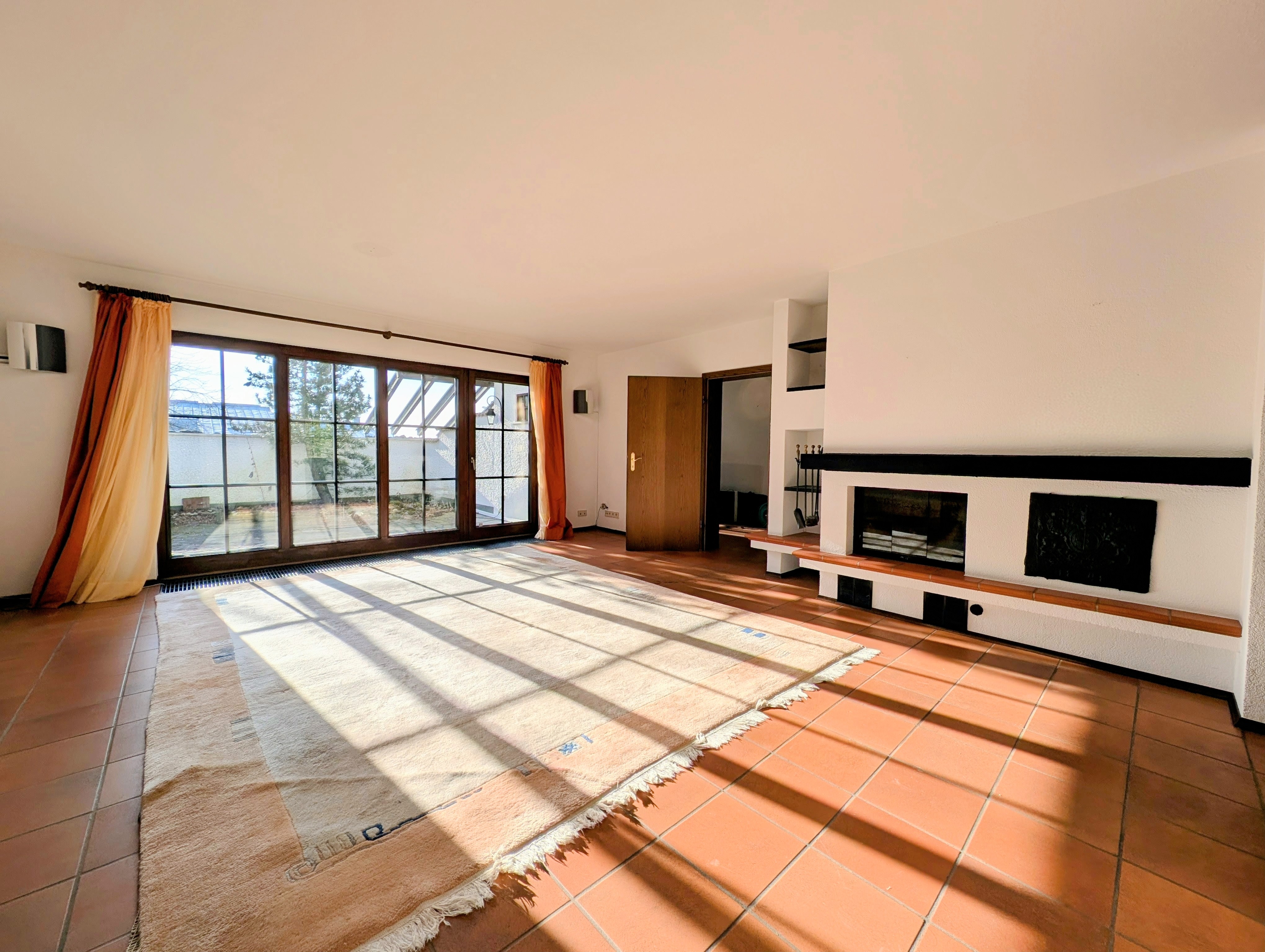 Bright living room with terracotta tile floor, large rug, fireplace, and sliding glass doors with orange curtains.