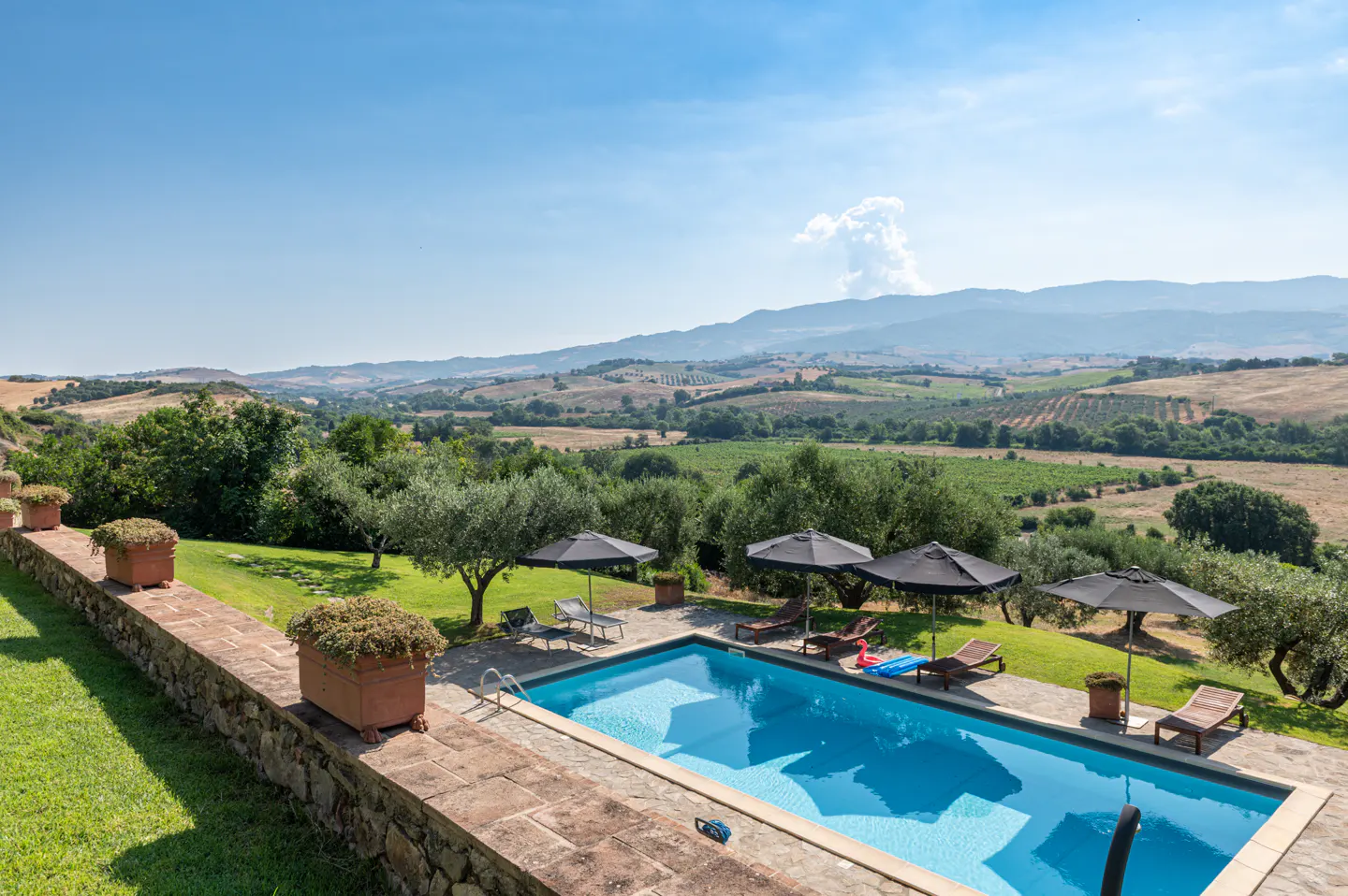A landscape view of a blue swimming pool with lounge chairs and umbrellas overlooking green hills and a blue sky.