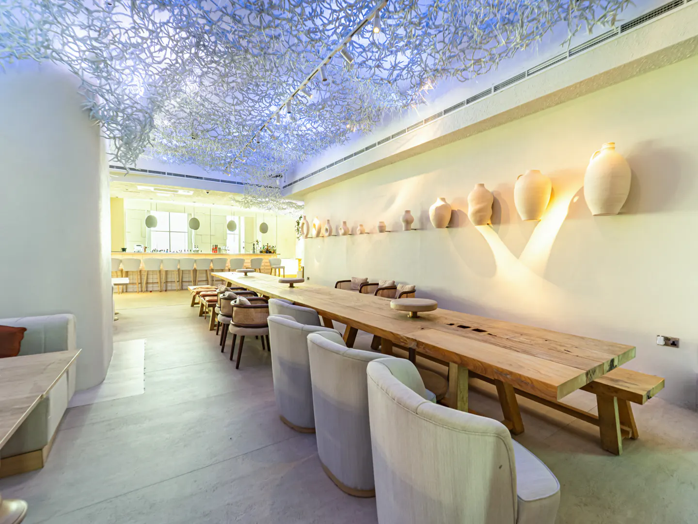 Modern restaurant interior with a long wooden table, upholstered chairs, and decorative clay vases on a cream-colored wall.